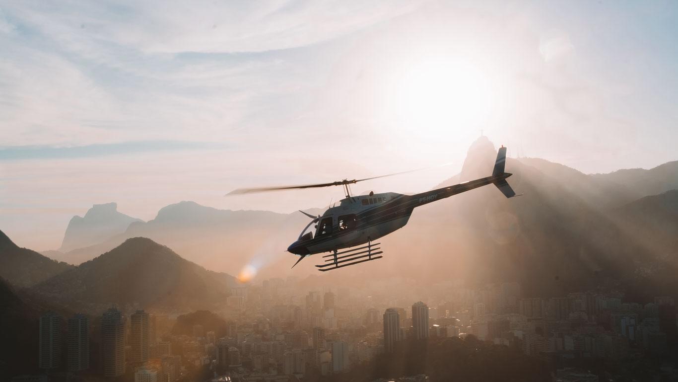 Un helicóptero sobrevuela la ciudad de  Río de Janeiro en enero, al atardecer, con montañas brumosas y el Cristo Redentor al fondo. La luz dorada del sol baña la escena, creando un ambiente cálido y aventurero.