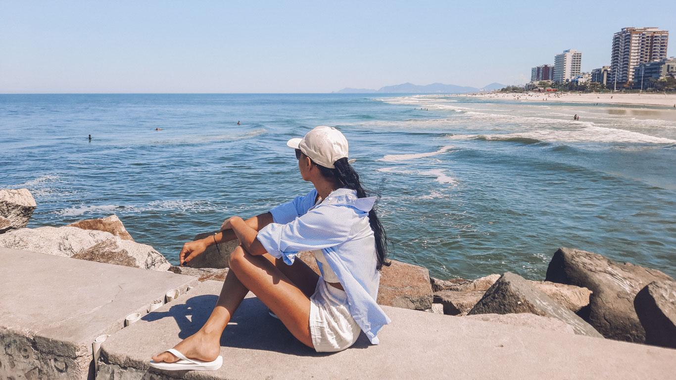 Mujer sentada en una escollera observa el océano en Barra da Tijuca, vestida con gorra, camisa holgada y sandalias. A lo lejos, se aprecian surfistas en el agua y una hilera de edificios junto a la playa en un día soleado.