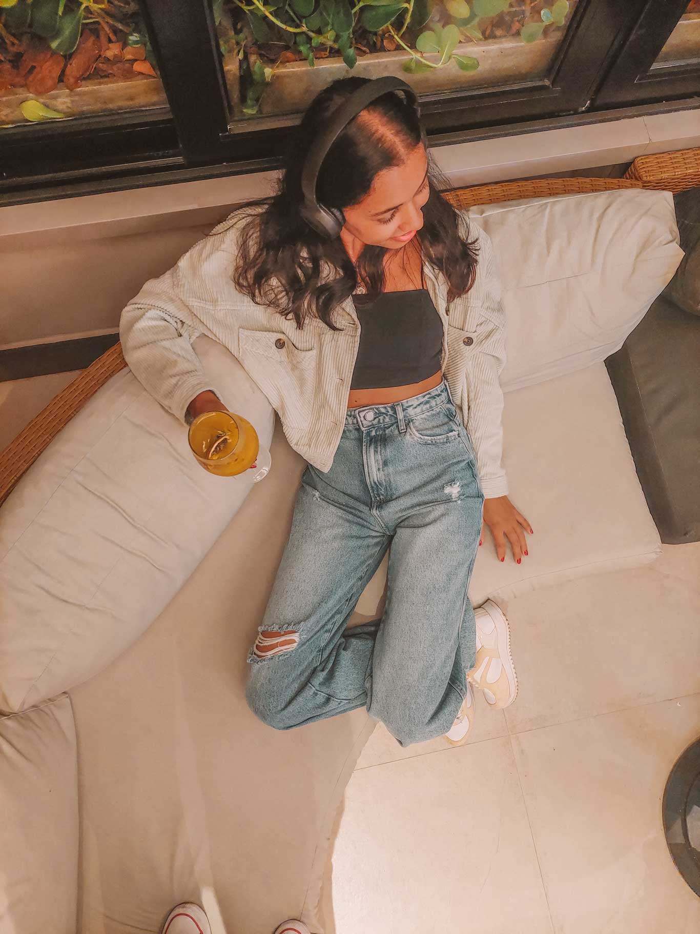 Chica sentada en un sofá beige del Mar Ipanema Hotel, uno de los mejores hoteles de Río de Janeiro, con auriculares negros, chaqueta clara, top negro y vaqueros rotos. Sostiene una bebida amarilla con una rodaja de fruta mientras se relaja en un espacio interior con plantas al fondo.