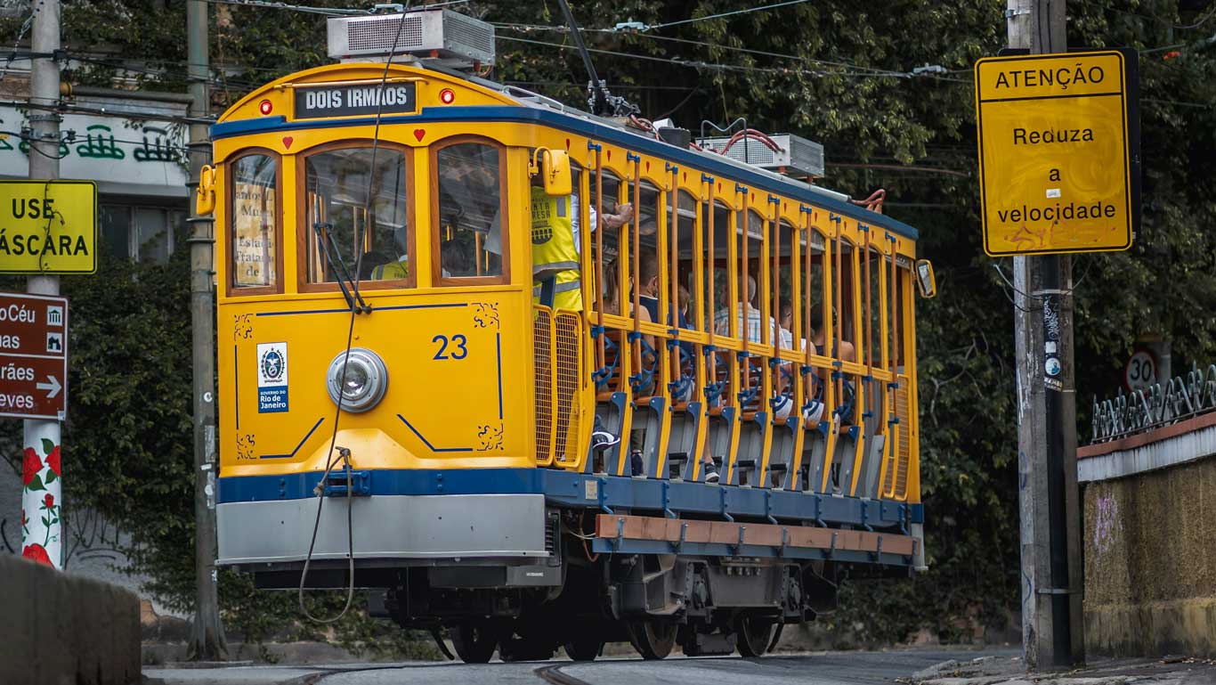 Tranvía amarillo tradicional del barrio de Santa Teresa, en Río de Janeiro, circulando por una calle con letreros de advertencia en portugués. El tranvía está lleno de pasajeros y mantiene su diseño clásico con ventanas abiertas y detalles azules.